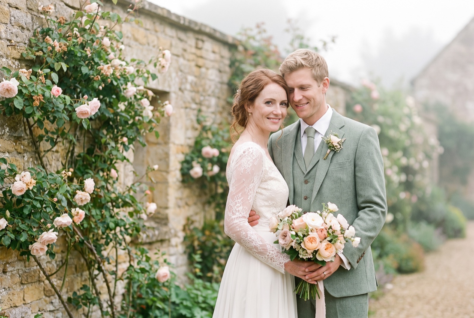 Eleanor and Frederick stand close together outdoors in soft summer light, foreheads nearly touching, both smiling gently.
